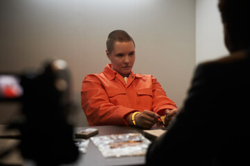 Caucasian young adult woman with short hair sitting at interrogation table in orange prison uniform...