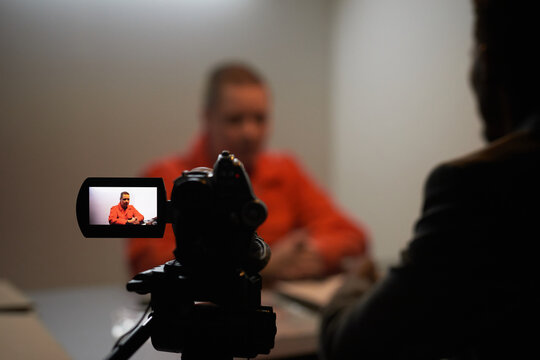 Caucasian middle aged woman wearing prison uniform sitting at table being filmed during interview with unidentified , camera screen showing subject in focus, prisoned context