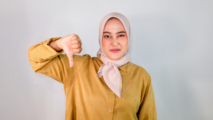 Muslim woman wearing hijab and brown shirt looking sad and disappointed, showing thumbs down and...