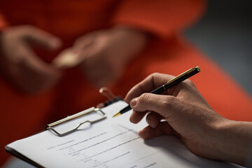 Caucasian woman holding pen and clipboard conducting interview with prisoned adult wearing orange...