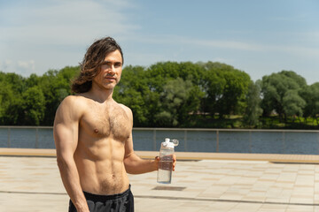 Shirtless handsome fit young man with long hair holding water bottle while standing outdoors on...