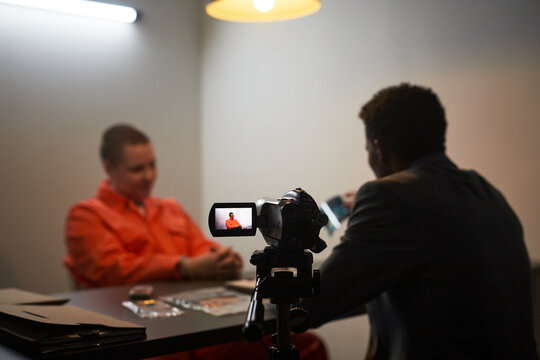 Caucasian middle aged man in orange prison uniform sitting at table during interrogation, Black man in suit interviewing and recording conversation with video camera in dimly lit room