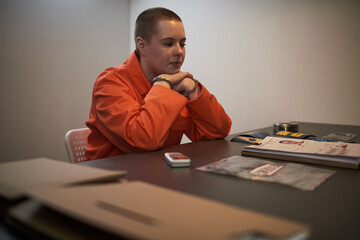 Caucasian young adult woman with short hair wearing orange prison uniform, sitting handcuffed at...