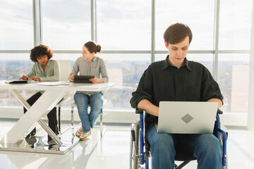 Accessible environment. Young man in chair for people with disability working with laptop in modern office. Diverse multiracial group of people colleagues working together. Young professional team