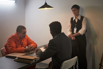 Caucasian young adult woman in orange prison uniform sitting at table with Black middle aged man in...