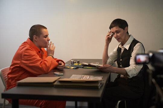Caucasian teenager with shaved head wearing orange prison jumpsuit, sitting at table smoking cigarette while middle aged Caucasian woman in formal attire reading documents during prison visit