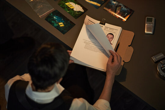 Middle aged Caucasian woman sitting at desk reviewing criminal case file with mugshot and evidence photographs, holding documents while analyzing investigation materials related to prisoned suspect