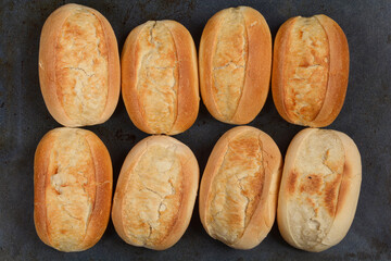Eight freshly baked, oval-shaped bread rolls are neatly arranged in two rows on a dark surface,...