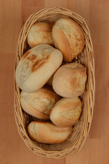 A rustic woven basket is filled with an assortment of freshly baked, golden-brown bread rolls, viewed from directly above on a wooden surface