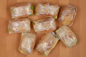 Several individual sandwiches, each wrapped in clear plastic film, are scattered across a light wooden surface, ready for a picnic or packed lunch