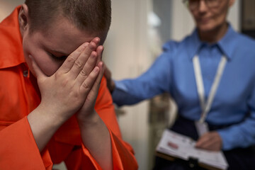 Caucasian teenager with short hair sitting with head in hands wearing prison uniform, showing...