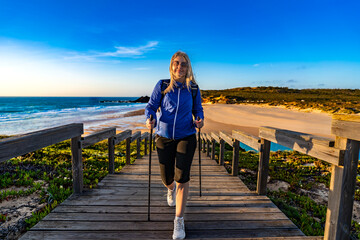 Beautiful mature woman practicing nordic walking on wooden stairs with sandy beach and ocean in...
