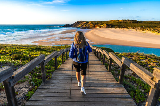 Beautiful mature woman practicing nordic walking on wooden stairs with sandy beach and ocean in background. Back view. Amoreira Praia on Algarve coast, Portugal		