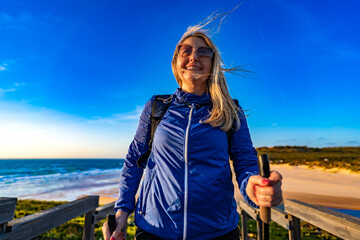 Beautiful mature woman practicing nordic walking on boardwalk with sandy beach and ocean in background. Front view. Amoreira Praia on Algarve coast, Portugal	
