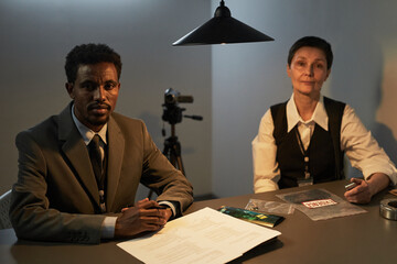 Black young adult man sitting at interrogation table with documents and evidence bags, Caucasian...