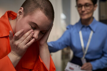 Caucasian young adult woman wearing prison uniform sitting with head in hands, showing distress while middle aged Caucasian woman in uniform standing nearby offering support