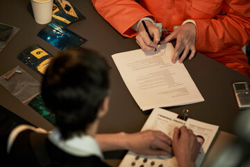 Caucasian woman in orange prison jumpsuit handcuffed, signing legal documents at table with...