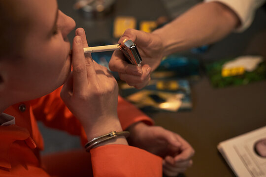 Caucasian young adult woman wearing orange prison uniform and handcuffs smoking cigarette while another lighting it, prisoned context with identification documents visible