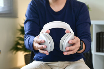 A man is holding a pair of headphones in his hands. The man is sitting on a chair, and there are several books around him