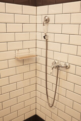 Close-up of clean modern shower corner with classic white subway tiles, chrome faucet, handheld showerhead and minimalist metal shelf. The interior details of the modern hotel in retro vintage style