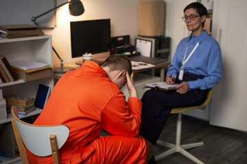 Caucasian young adult woman wearing prison uniform, sitting with head in hands in front of middle...