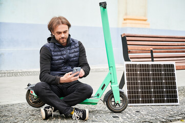 Man sits on electric scooter with solar panel and smartphone while charging. Integration of...