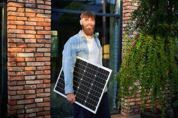 Portrait of bearded man holds solar panel, standing in front of brick building and greenery....