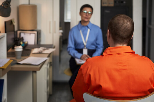 Caucasian young adult woman in orange prison uniform sitting with back to camera, facing middle aged Caucasian woman in professional attire conducting interview in prison office setting