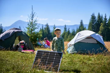 Fotobehang Young child playing near tourist tent with photovoltaic solar panel in summer. On background of mountains and trees under clear blue sky. Integration of renewable energy in outdoor camping activities. © anatoliy_gleb