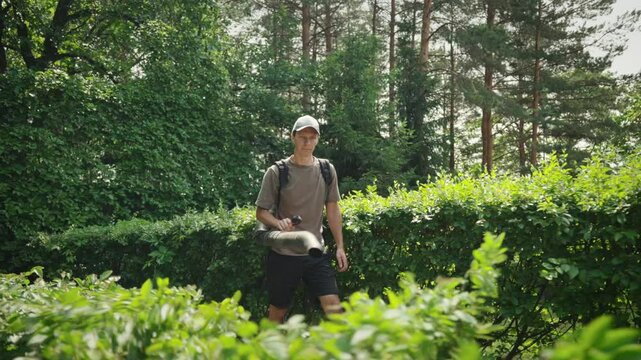 A man wearing a cap and casual clothes is using a backpack leaf blower in a lush, green garden. He is surrounded by neatly trimmed bushes and tall trees. 
