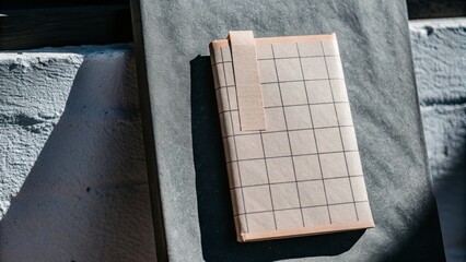 A checkered notebook with a beige tab rests on a dark grey fabric surface with textured wall shadows in the background.
