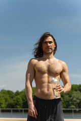 Brunette topless man with curly long hair holding water bottle looking at camera against clear blue sky and nature background. Heat summer.