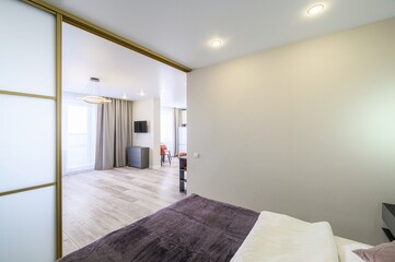 Modern apartment view with a bedroom section.  A partly visible bed dominates the foreground, flowing into a sleek living area with muted colors