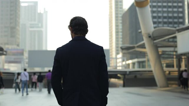 Businessman walks across urban square at modern downtown block backside view. Company CEO young man strolls observing stylish architectural forms of megapolis. Business success