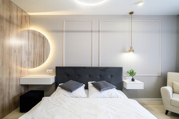 modern bedroom with a grey headboard, white linens, and a round, illuminated mirror above a vanity