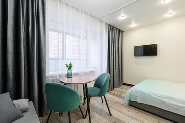 bright studio apartment features a small dining area near a window with green chairs and a bed with a TV on the wall