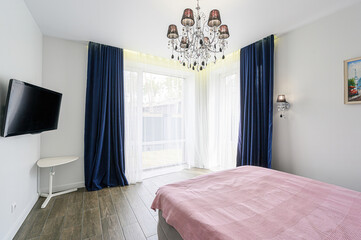 bright bedroom featuring a bed with a pink cover, large windows with blue curtains, a chandelier,...