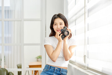 Young Asian Woman Photographer in White T-Shirt Holding Vintage Camera by Bright Window Indoors	
