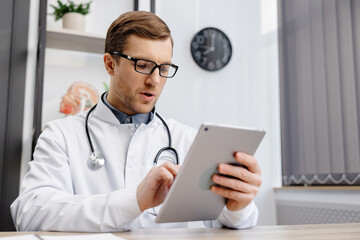 Male Caucasian doctor in white medical uniform filling patient medical history or anamnesis on tablet in office. Doctor consulting client online on tablet in modern clinic, healthcare concept