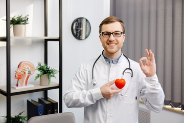 Doctor in white coat and glasses holding a red heart at hospital office. Medical health care and...