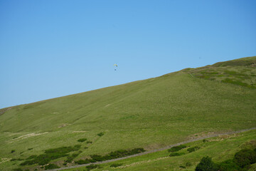 Paraglider flying over rolling green hill under clear blue sky in England, a popular paragliding...