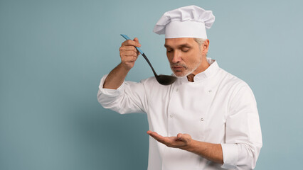 Mature male chef with spoon tasting dish on light blue background.