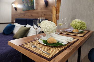 A beautifully arranged dining table features a vase of white flowers, wine glasses, and a bread roll, all set in an inviting hotel room with wood accents and soft lighting.