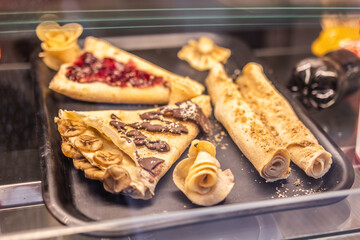 Assorted sweet crepes with chocolate, fruit, and crumbles on black tray. Street food, dessert culture, and indulgent culinary variety in cozy cafes.