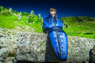 Man in a sleeping bag sitting on a large rock in the mountains on a sunny day. Outdoor adventure,...