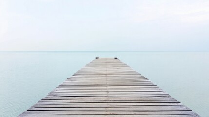 Fototapeta premium Weathered wooden pier extending into calm light-blue sea with gentle ripples, small black mooring posts at end, pale sky with wispy clouds, serene horizon.