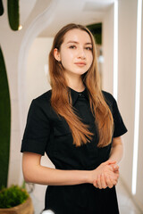 Professional female cosmetologist wearing black uniform looking at camera, standing confidently...