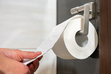 Close-up of hand unrolling toilet paper from dispenser on tiled wall, representing hygiene,...