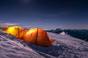 Illuminated orange tents on snowy mountain slope under starry night sky. Winter camping during...