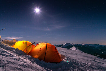 Illuminated orange tents on snowy mountain slope under starry night sky. Winter camping during...